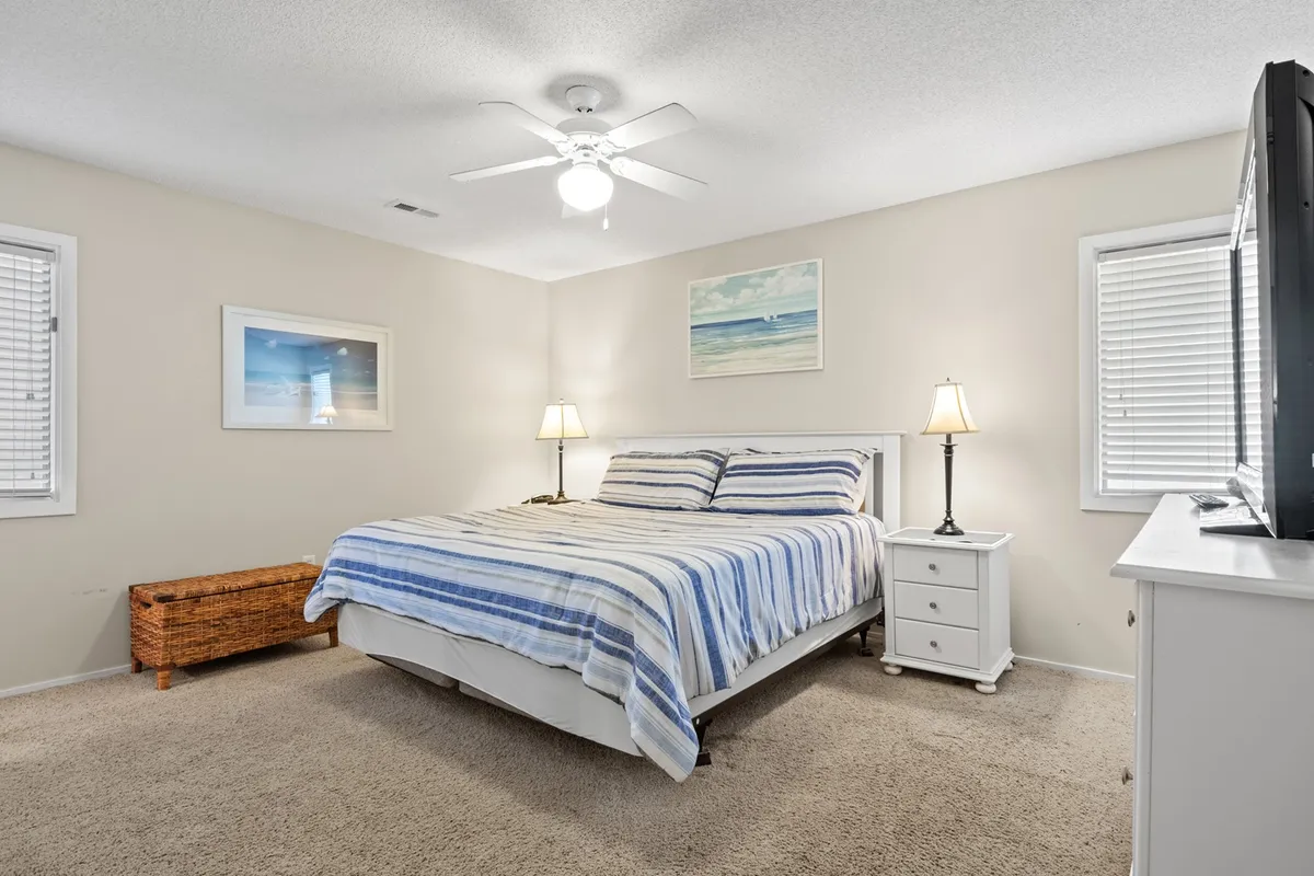 This image features a cozy bedroom with a bed adorned in a striped comforter, flanked by nightstands with lamps. Artwork decorates the wall, and a television is visible, offering all the comforts one would expect in a private retreat. Natural light spills into the room, enhancing its welcoming atmosphere.