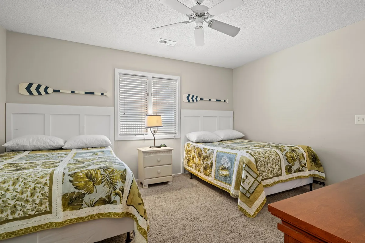 A bedroom featuring two beds with patterned bedspreads, a nightstand with a lamp, and a window with blinds. Decorative oars on the wall add a playful coastal touch. The room is softly lit and encourages socializing or quiet relaxation.