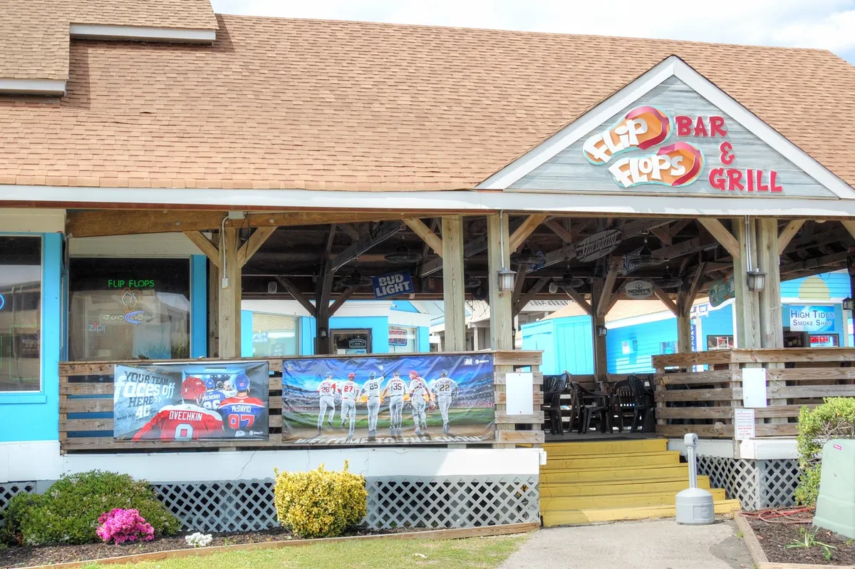 An inviting exterior view of Flip Flops Bar & Grill, featuring colorful banners and an outdoor seating area where guests are enjoying food and drinks. The relaxed setting suggests a perfect place for casual dining or socializing after a day of shopping or exploring.