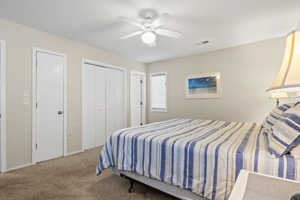 A peaceful bedroom with a striped bedspread, ceiling fan, and framed artwork. Windows allow sunlight to gently fill the space, creating a calming and serene mood, perfect for rejuvenation and restful sleep.