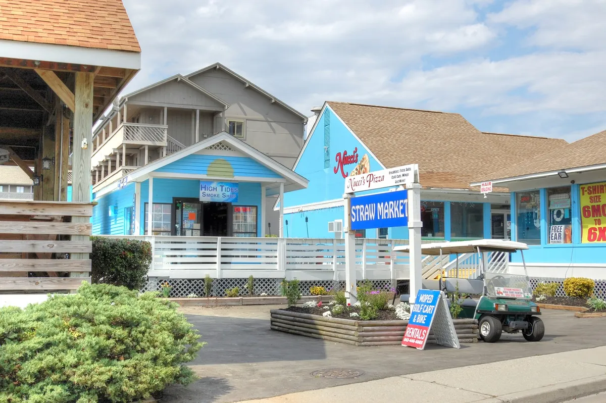 A welcoming exterior view of colorful blue buildings with clear signage and a golf cart parked outside, suggesting easy access to local shops and eateries. The lively scene hints at the proximity of markets, pizza places, and other attractions, all within a cheerful coastal environment.