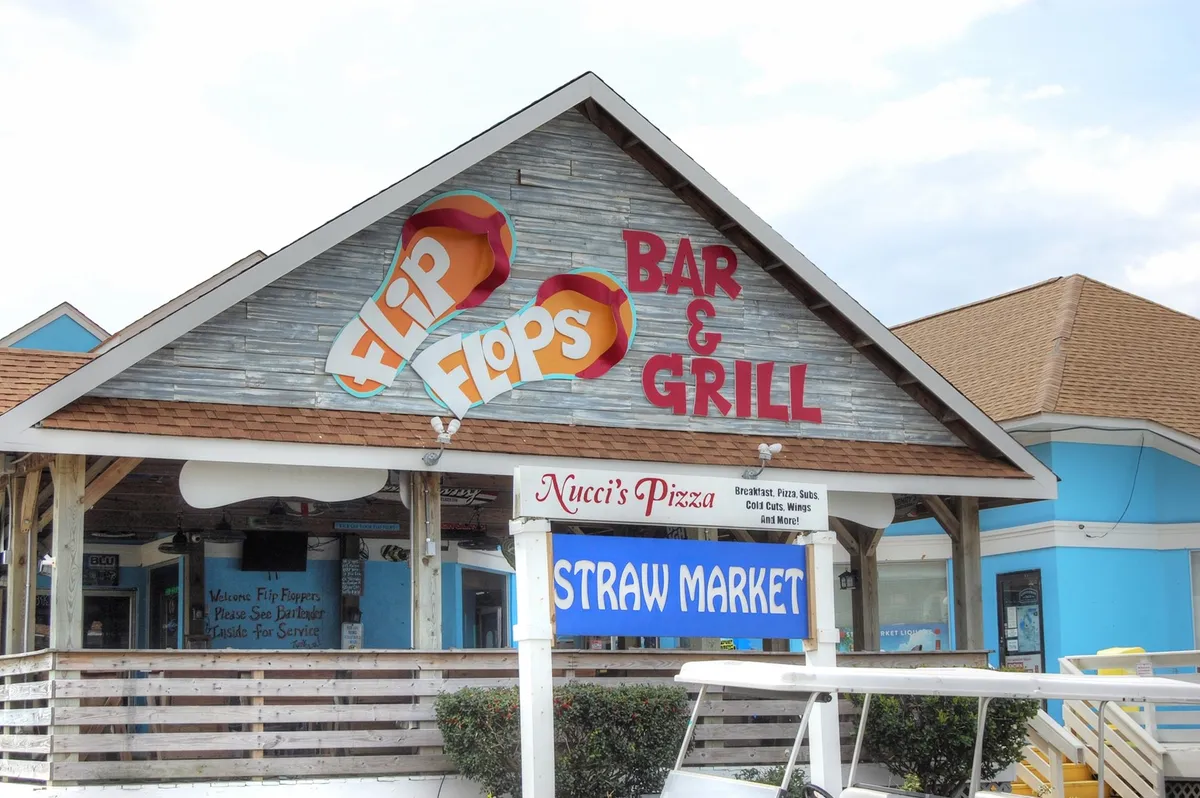 This exterior shot captures the front signage of Flip Flops Bar & Grill, Nucci's Pizza, and the Straw Market, all set against a backdrop of bright colors and inviting storefronts. People can be seen milling about, adding a sense of vibrant activity and local charm to the scene.
