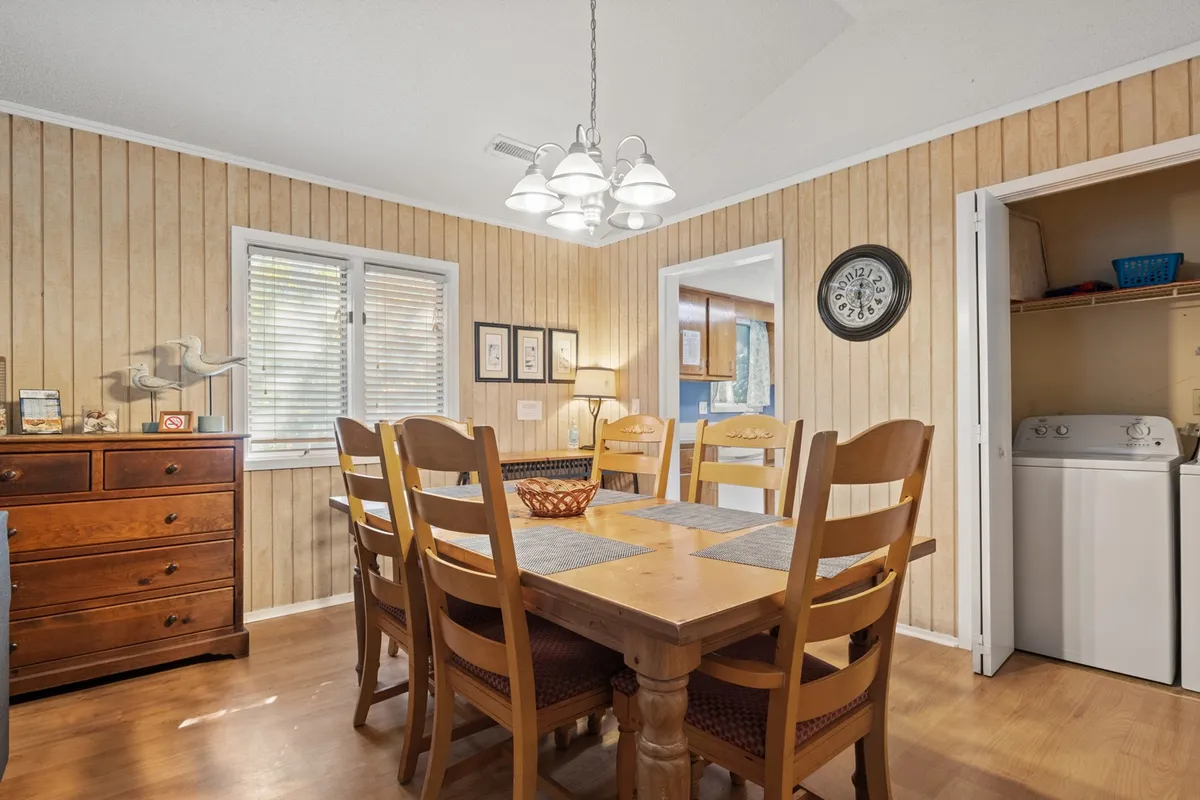 A welcoming dining area with a wooden table and chairs, a graceful chandelier overhead, and a window with blinds. The setting exudes warmth, perfect for family dinners, game nights, or gatherings with friends, seamlessly extending from the living space.