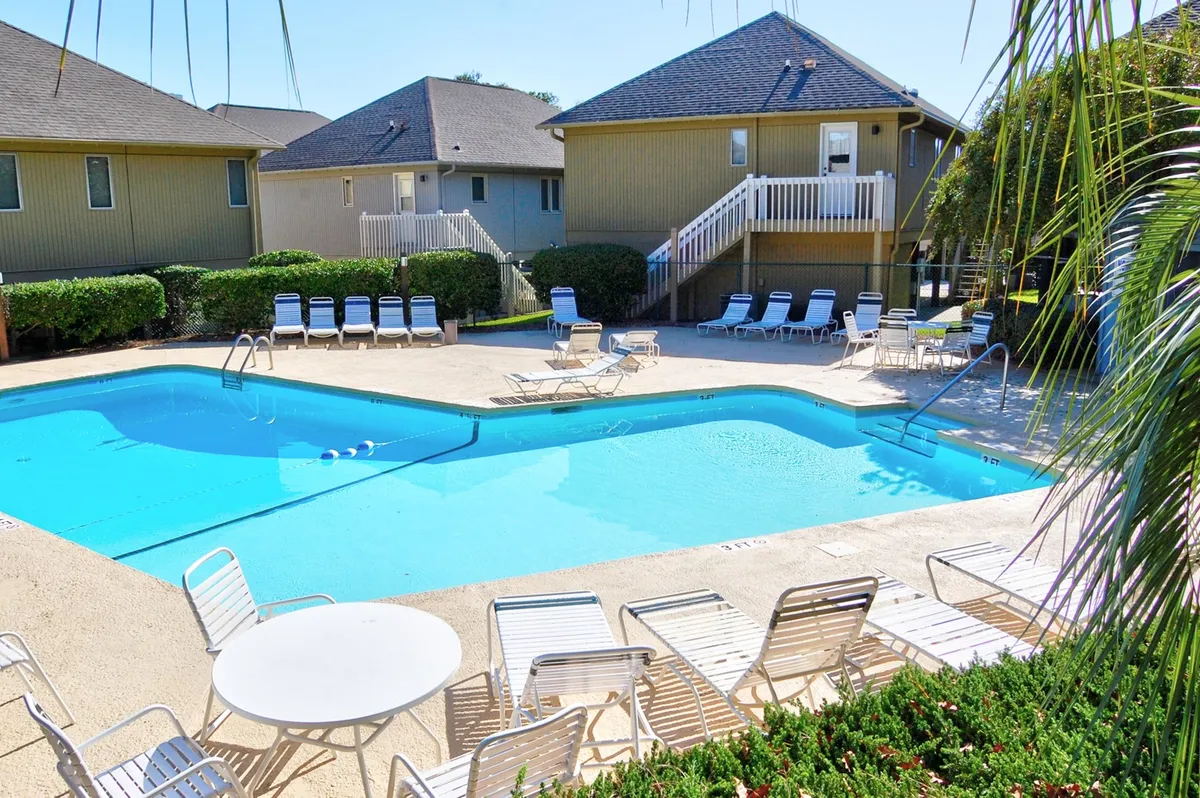 A vibrant community pool area lined with lounge chairs, surrounded by lush greenery and set against charming resort buildings. The atmosphere is leisurely, suggesting fun and relaxation for guests of all ages.
