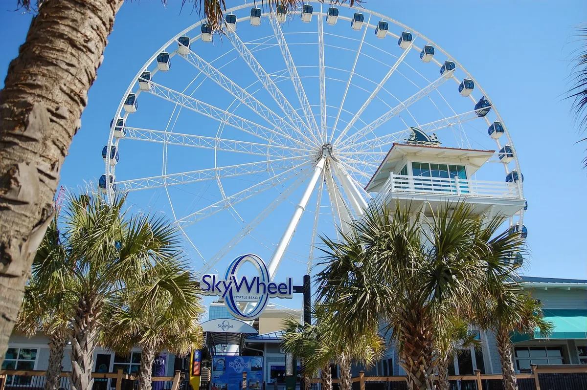 This image features the iconic Myrtle Beach SkyWheel rising above a backdrop of swaying palm trees and the bustling ticket booth, set against a bright, inviting sky. The scene captures the excitement and energy of one of Myrtle Beach’s most beloved attractions, with visitors lining up to enjoy panoramic views of the coastline.