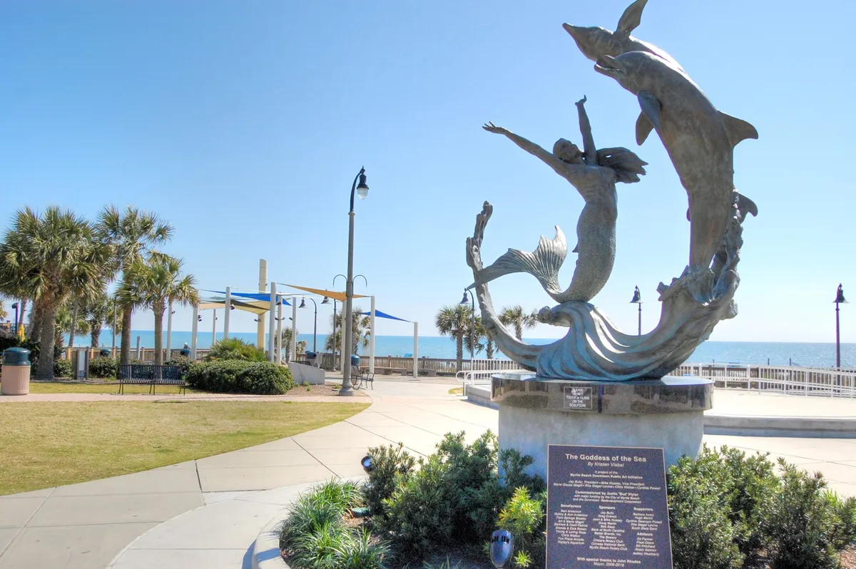 A serene scene on the Myrtle Beach oceanfront, featuring a striking statue of the Goddess of the Sea, palm trees, and a pathway leading toward the water. The image blends natural coastal beauty with local art, inviting viewers to imagine leisurely strolls and beachside adventures.