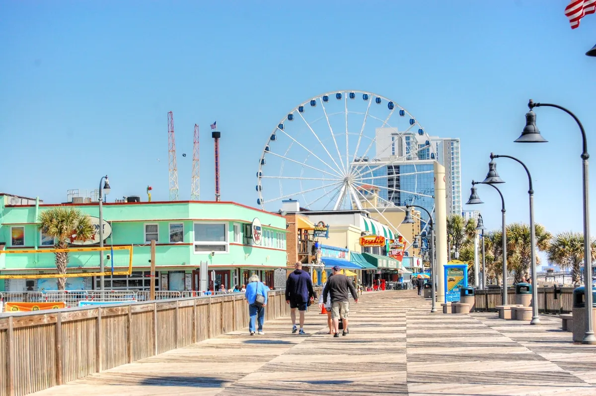A lively, colorful scene along the Myrtle Beach boardwalk featuring the Ferris wheel, palm trees, vibrant buildings, and people enjoying walks, shopping, and rides under a bright sky.