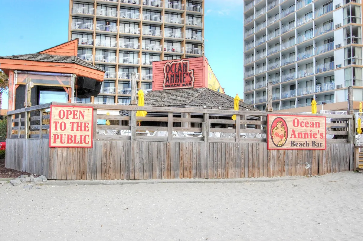 A laid-back exterior scene featuring the Ocean Annie's Beach Bar sign, open-to-the-public signage, wooden barriers, and bright yellow umbrellas. The area hints at a lively, welcoming beachfront setting, with opportunities to relax and socialize just steps from the sand.