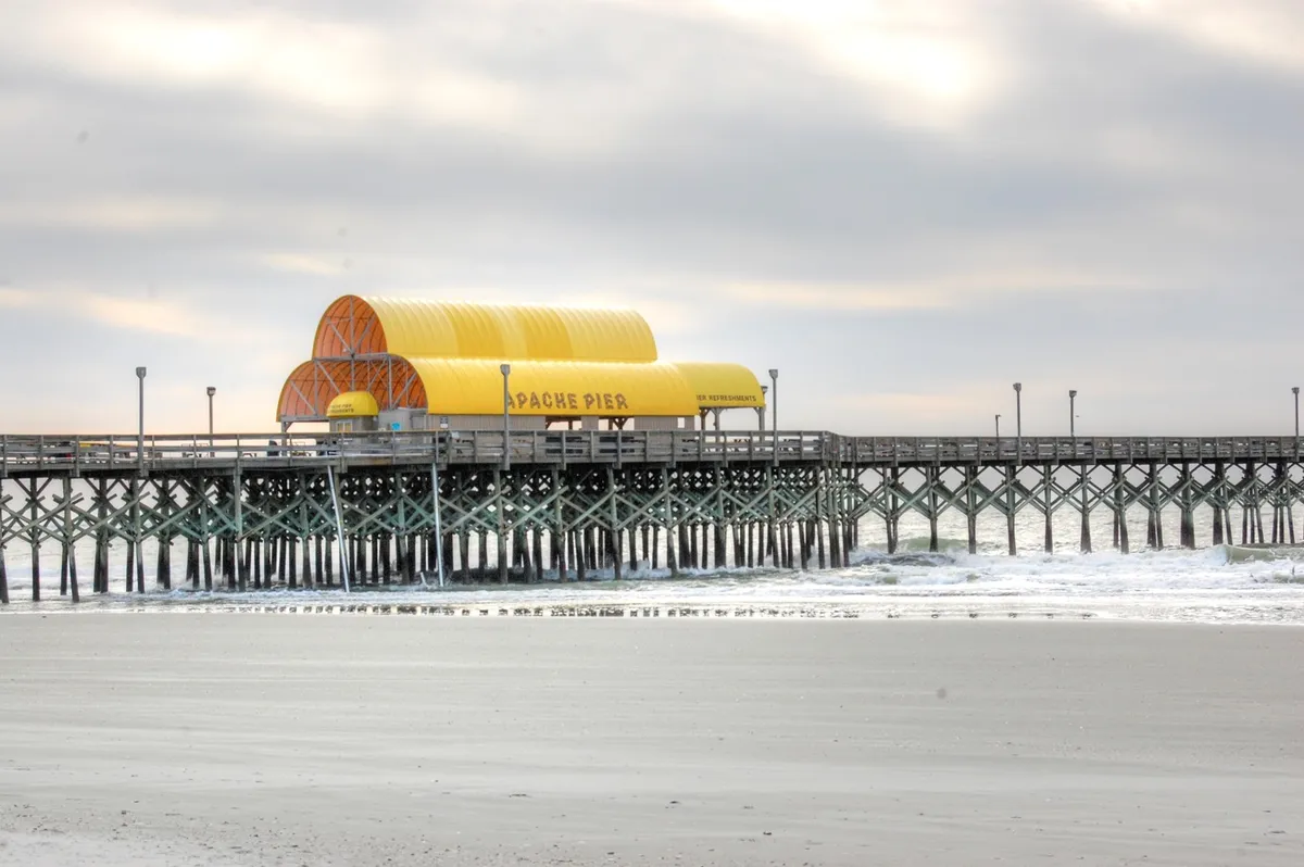 A view of the iconic Apache Pier with its bright yellow roof stretching out over the ocean. The wooden structure is surrounded by glistening water, inviting visitors to stroll, fish, or take in scenic views of the coastline.