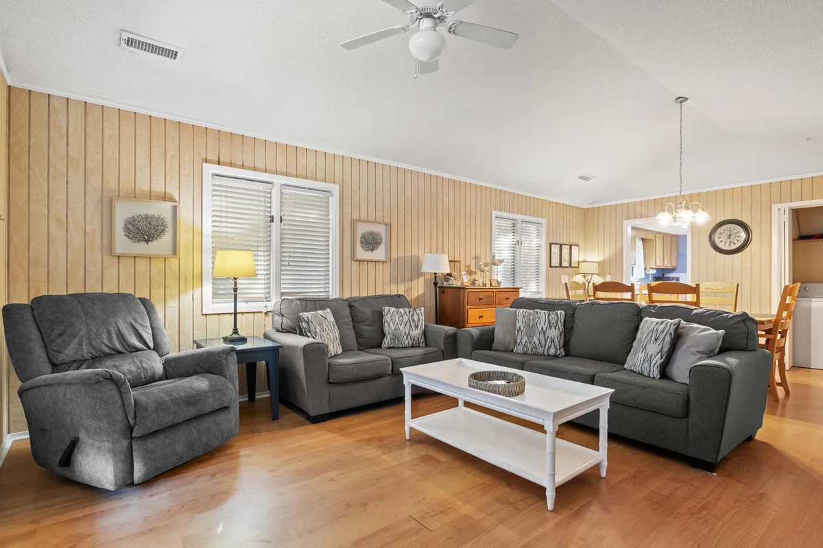 A spacious living room furnished with a plush sofa set, coffee table, and an inviting armchair, all illuminated by soft overhead lighting. The arrangement invites conversation and relaxation, perfect for gatherings or cozy evenings in. The space is open, comfortable, and designed for social connection.
